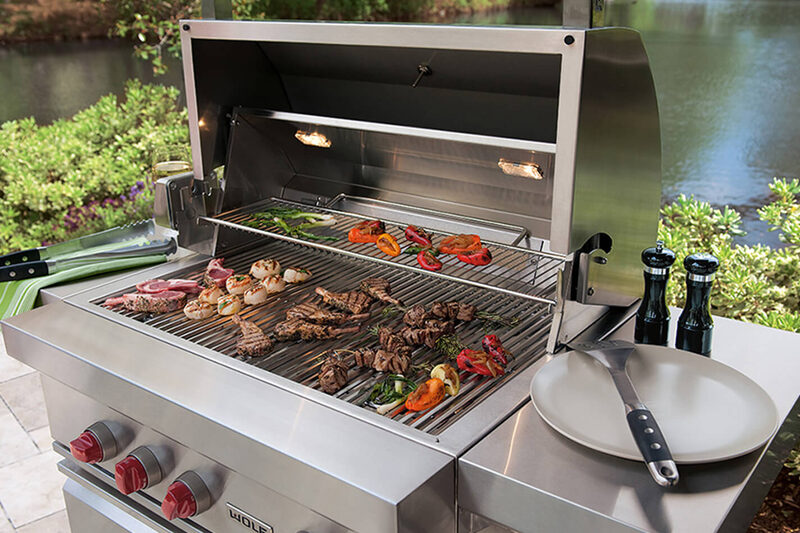 A built-in outdoor gas grill cooking steaks and vegetables beside a waterfront patio.