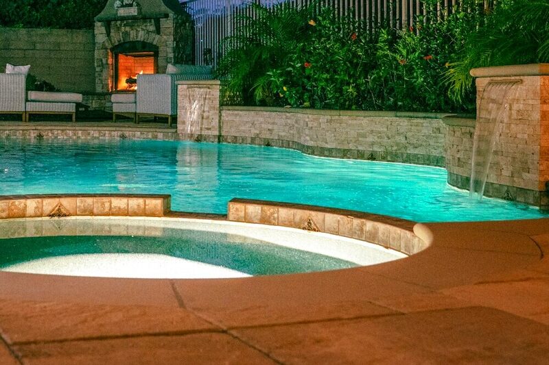 A lit pool and spa steaming at dusk beside an outdoor fireplace.