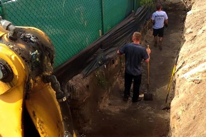 An Elite Gas crew installing a yellow propane gas line in an open trench.