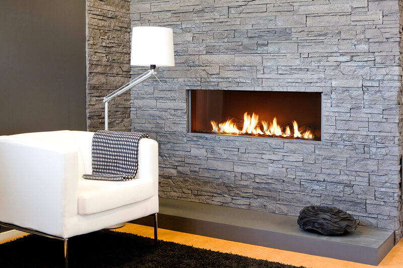 A modern linear gas fireplace set into a stacked stone wall beside a white armchair.